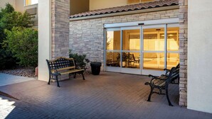 Un espace extérieur aménagé en patio, avec deux bancs, une plante en pot et l’entrée d’un bâtiment dotée d’une porte en verre.