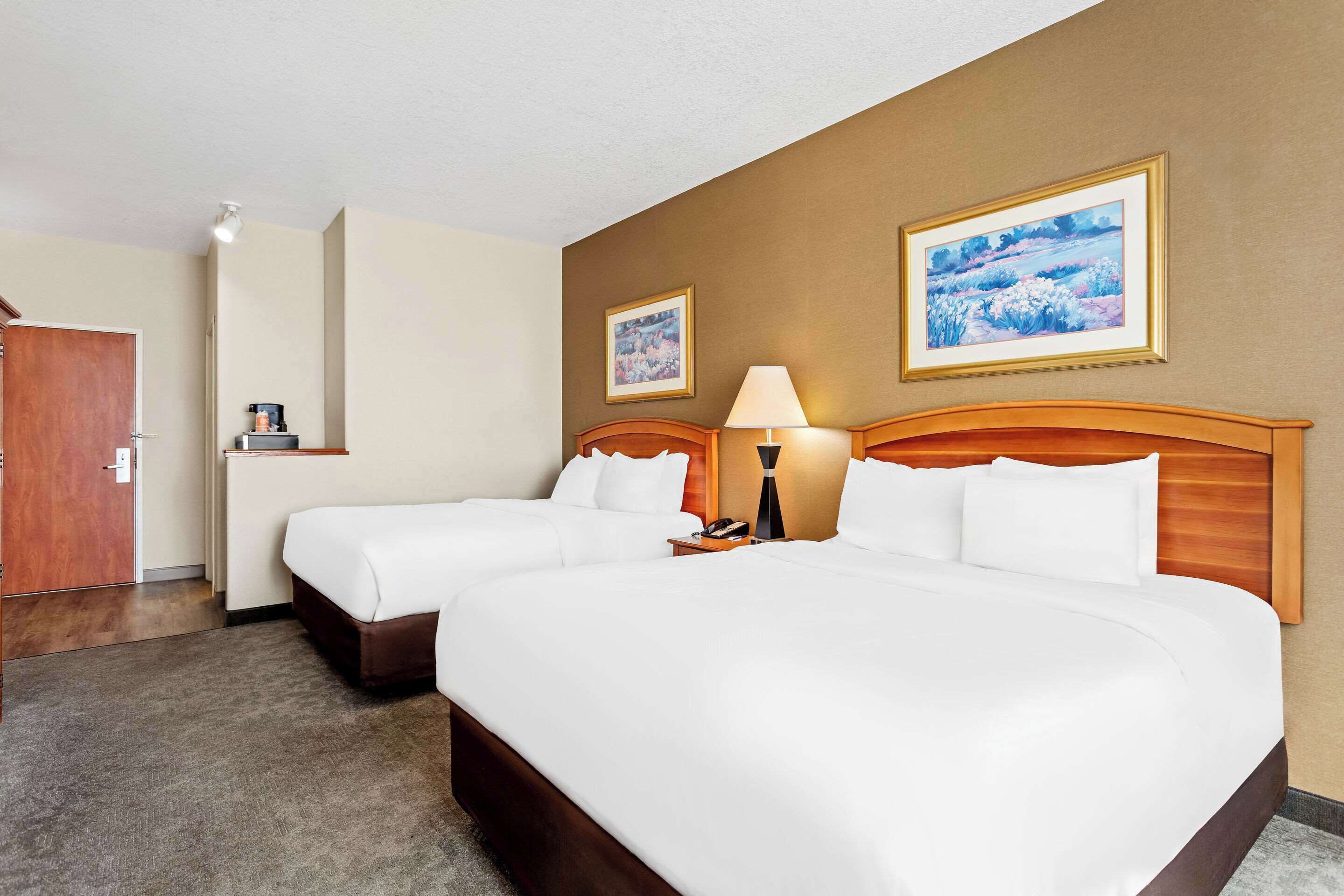 A hotel room with two beds, a wooden headboard, a bedside table with a lamp, and two framed pictures on the wall.