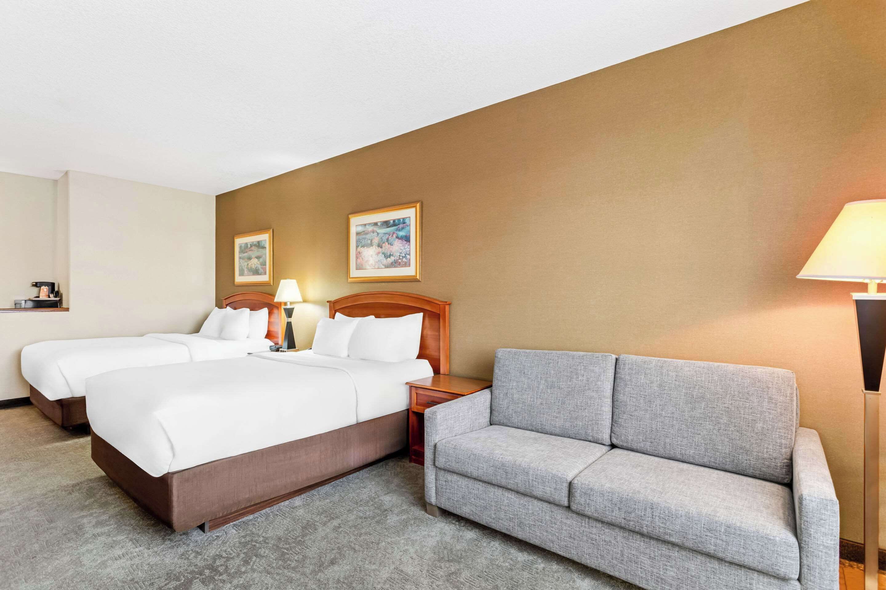 A hotel room with two beds, a grey sofa, a wooden headboard, and two framed pictures on the wall.