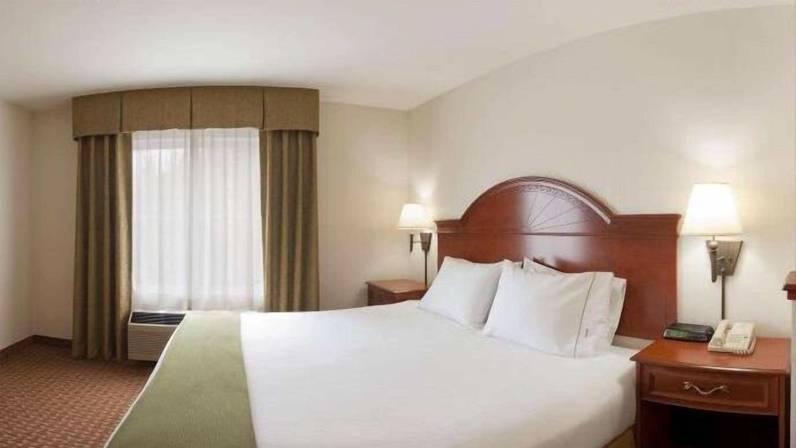 Hotellihuone, jossa on suuri sänky, kaksi yöpöytää, yölamppu, lamppu ja ikkuna, jossa on verhot.
