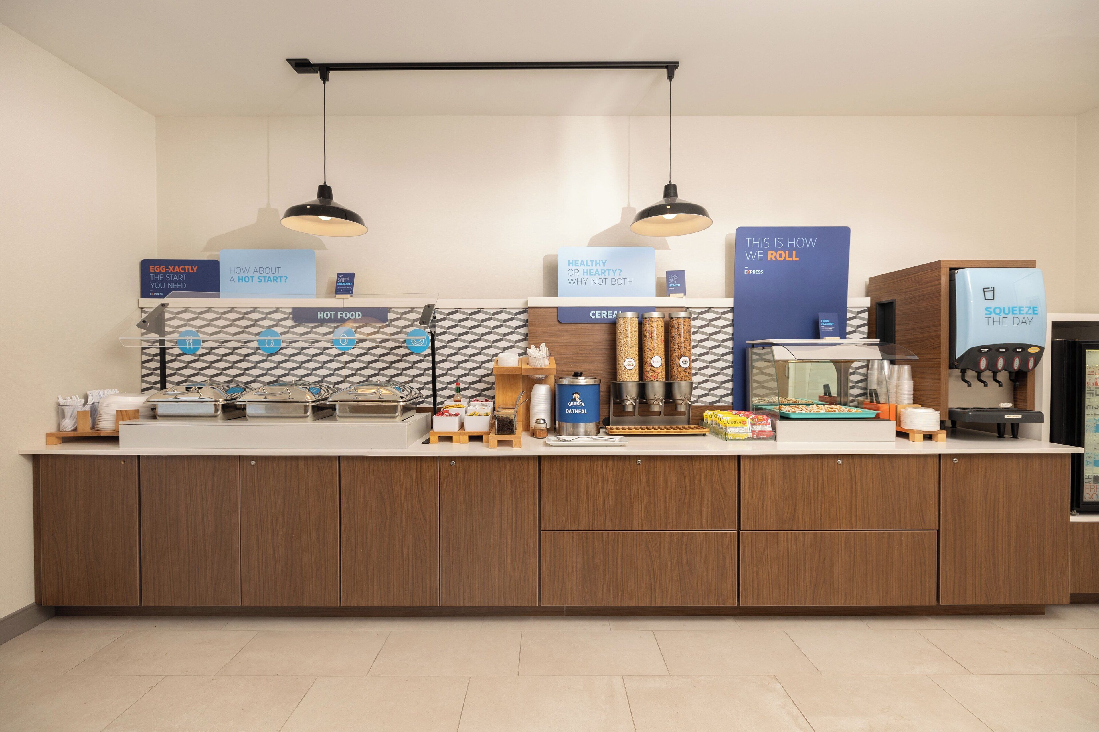 A modern kitchen area with wooden cabinets, a countertop with various food items and containers, and overhead pendant lighting.