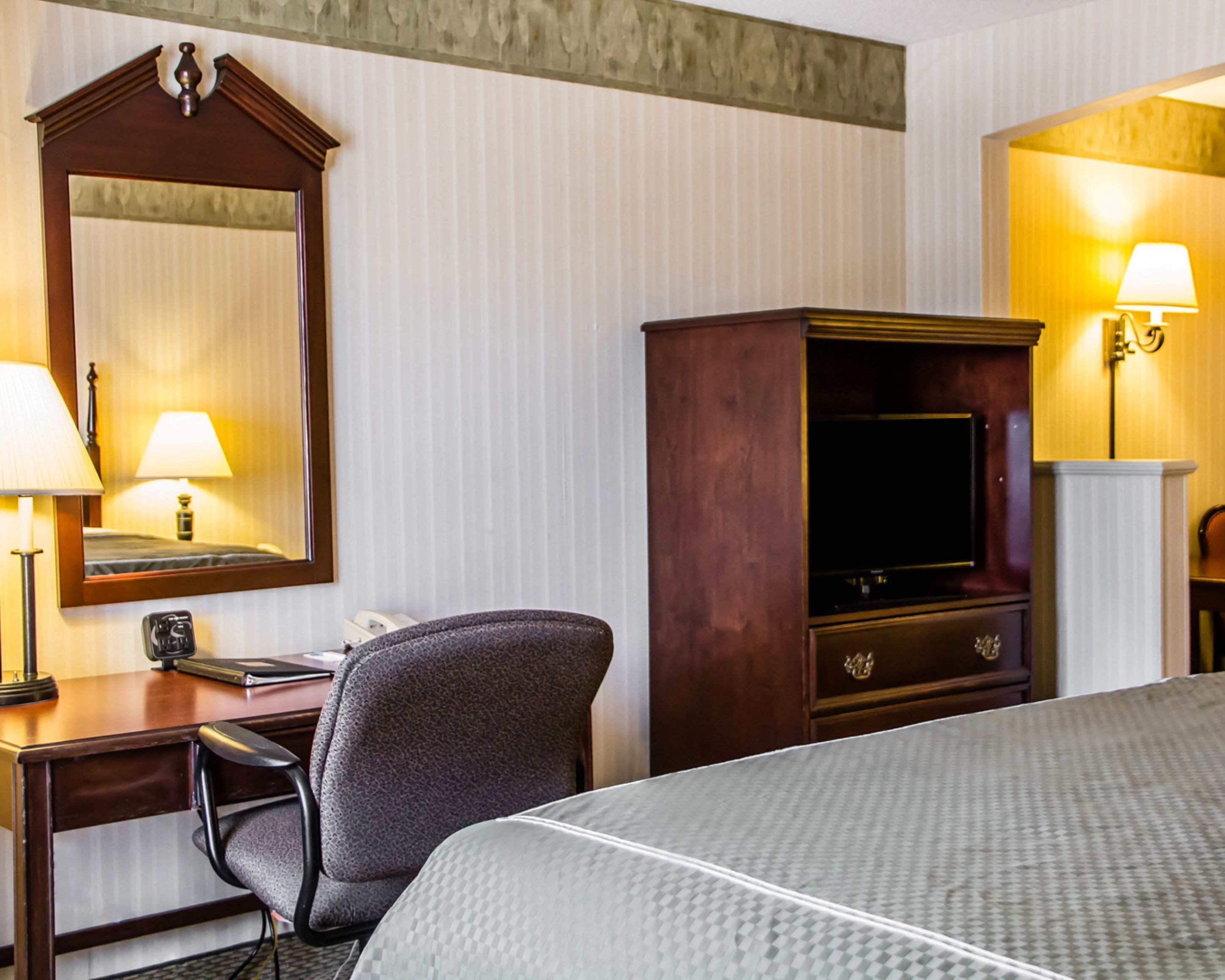 A hotel room with a bed, a desk with a chair, a television, a mirror, and two lamps.