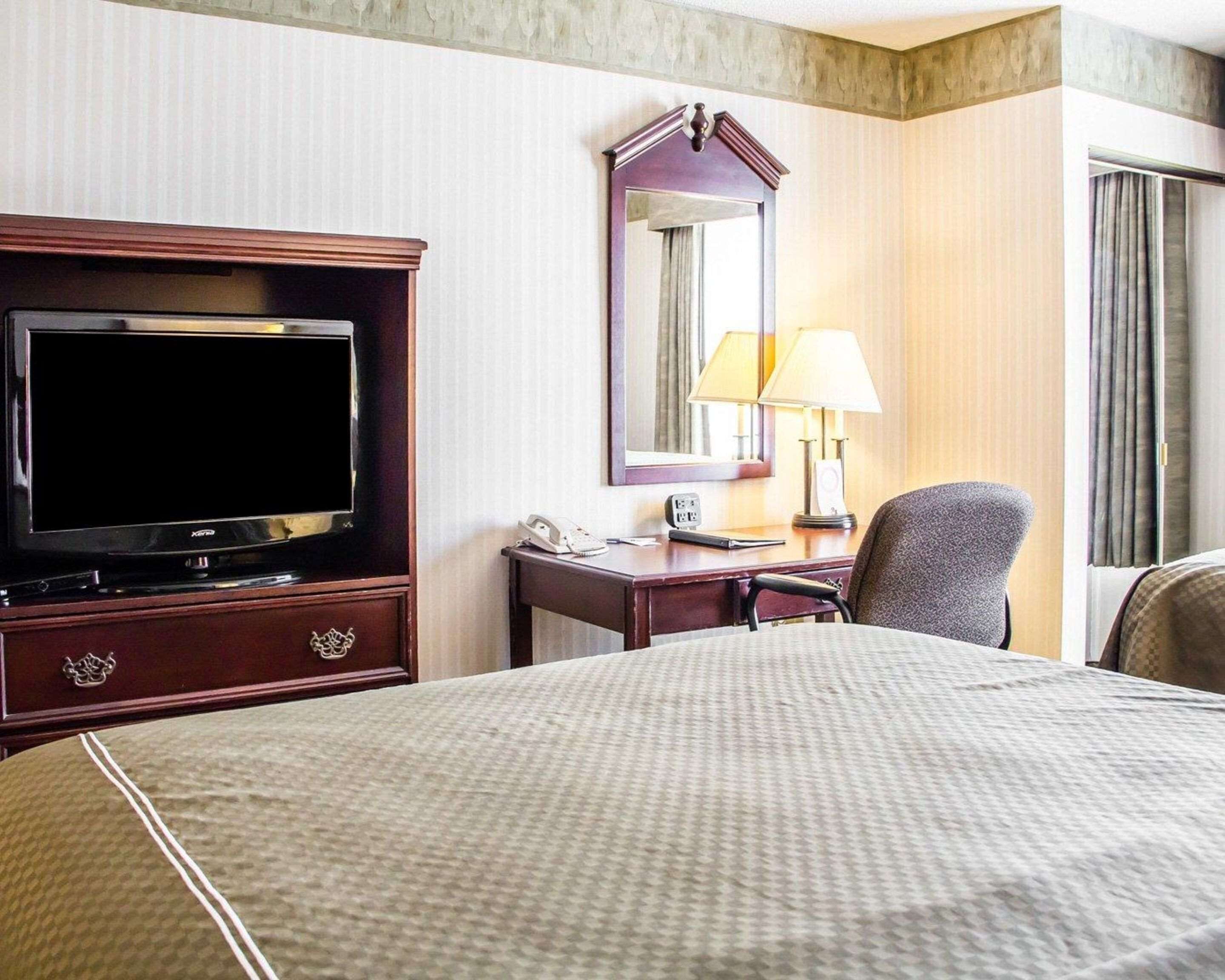 A hotel room with a large bed, a desk with a lamp, a television, and a mirror.