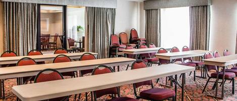Un salón de conferencias con largas mesas y sillas, ventanas amplias con cortinas y una alfombra estampada.