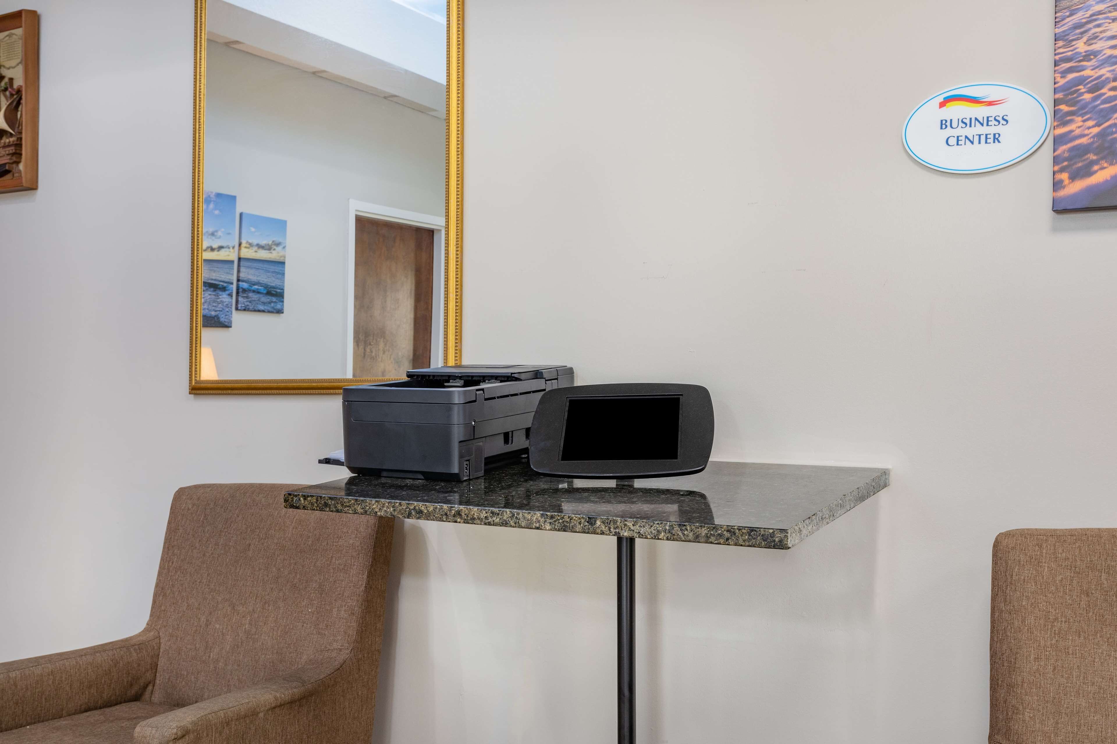 A reception desk with a printer, a computer monitor, and a mirror. There are two chairs and a wall-mounted sign that reads "BUSINESS CENTER".