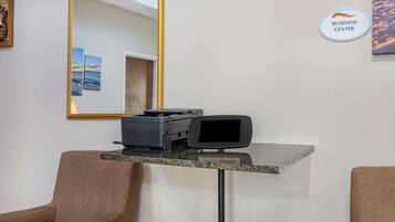 A reception desk with a printer, a computer monitor, and a mirror. There are two chairs and a wall-mounted sign that reads "BUSINESS CENTER".
