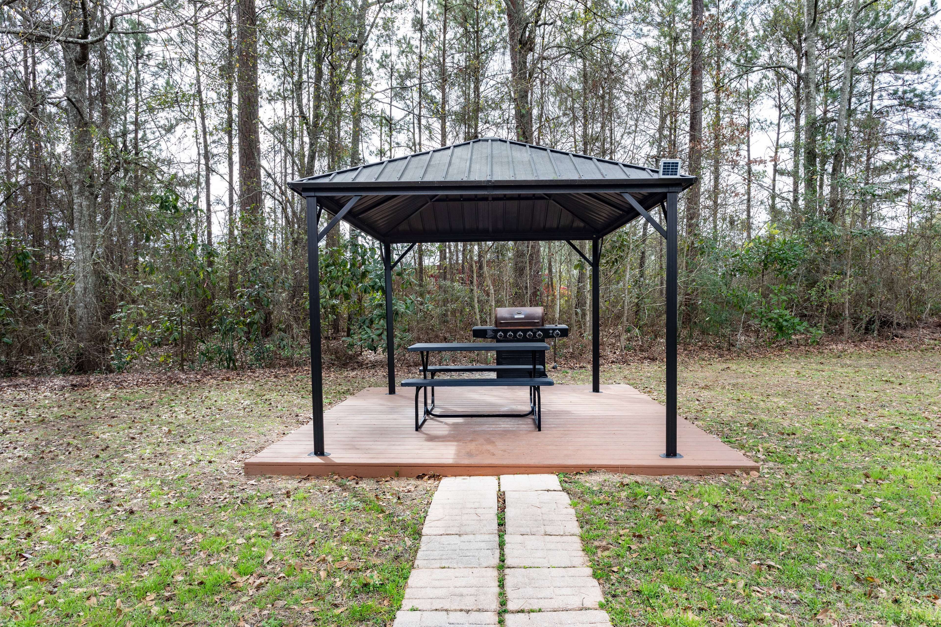 A covered outdoor area with a grill and picnic table, surrounded by a wooded area.