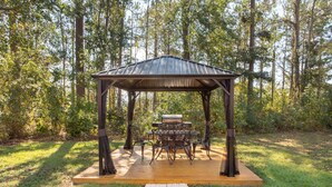 A gazebo with a table and chairs, surrounded by a wooden deck and a forested area.