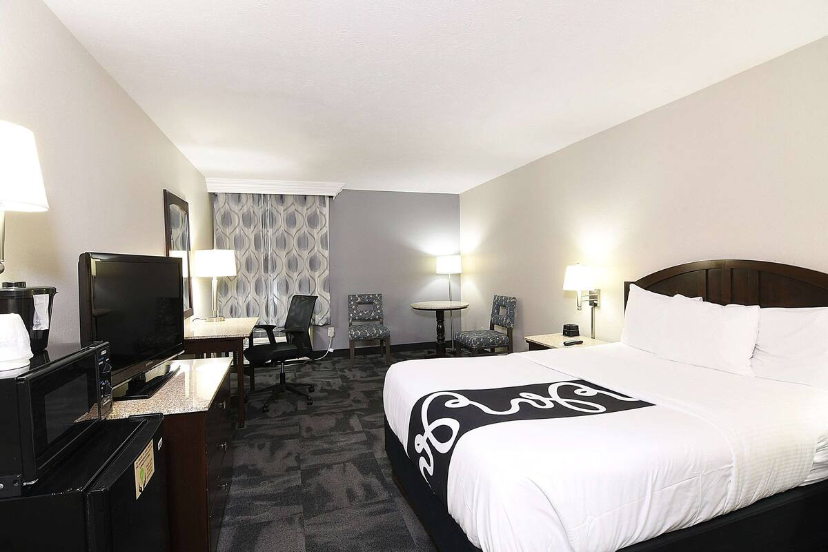 A hotel room with a bed, a desk with a computer, a TV, two chairs, a lamp, and a wall-mounted clock.