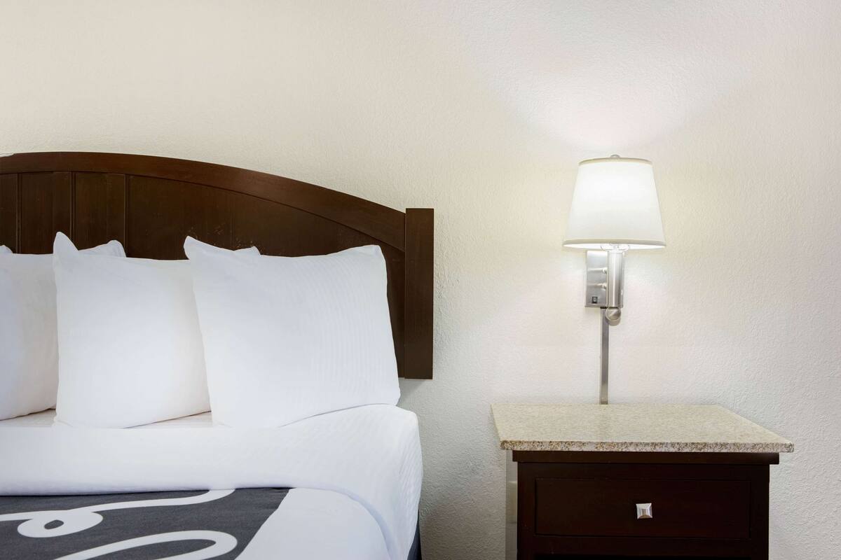 A neatly made bed with white linens and a headboard, a wall-mounted lamp, and a small wooden nightstand with a drawer.