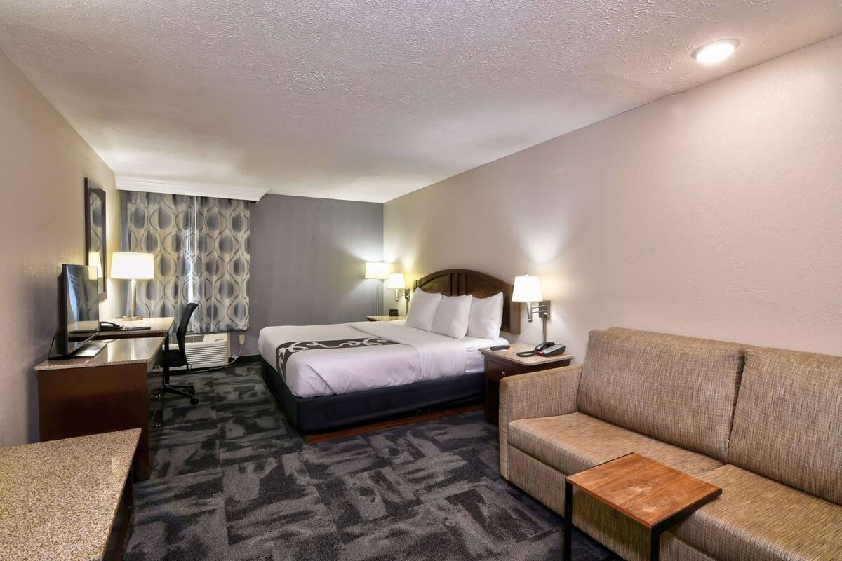 A hotel room with a bed, a desk, a chair, a sofa, a small table, and a lamp.