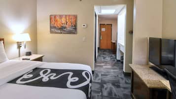 A hotel room with a bed, a TV, a painting, and a hallway leading to a bathroom.