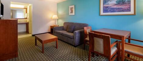 A hotel room with a sofa, a wooden table, a chair, a TV, a lamp, a framed picture, and a carpeted floor.