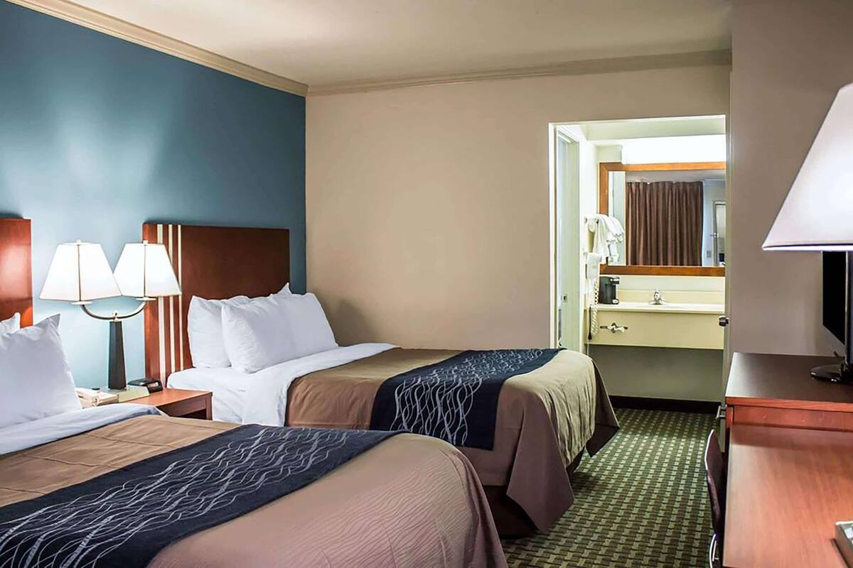 A hotel room with two beds, a desk, a chair, a lamp, a mirror, and a bathroom visible through an open door.