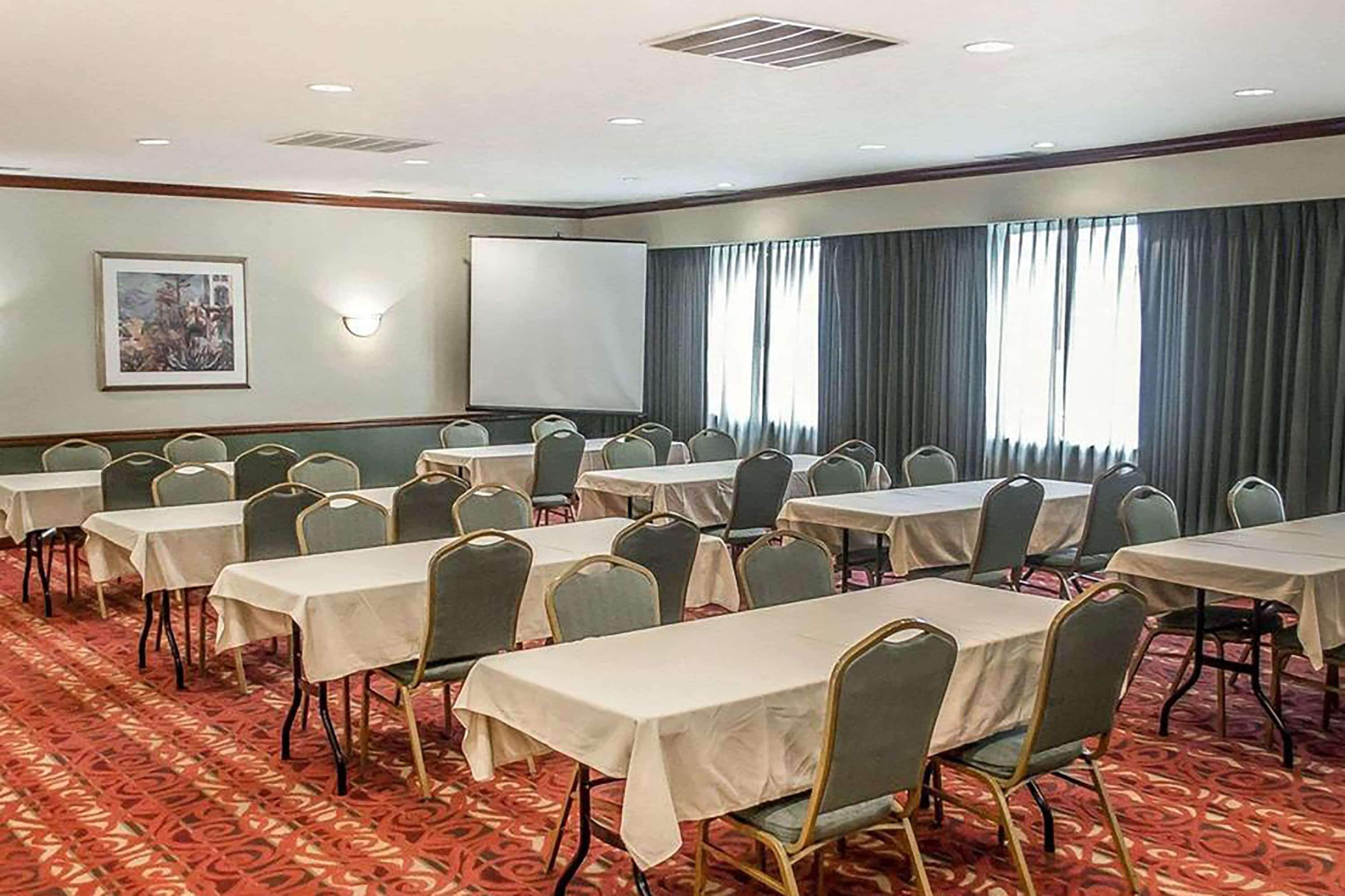 Un salón de conferencias con varias mesas y sillas, una pantalla de proyector y un amplio ventanal con cortinas.
