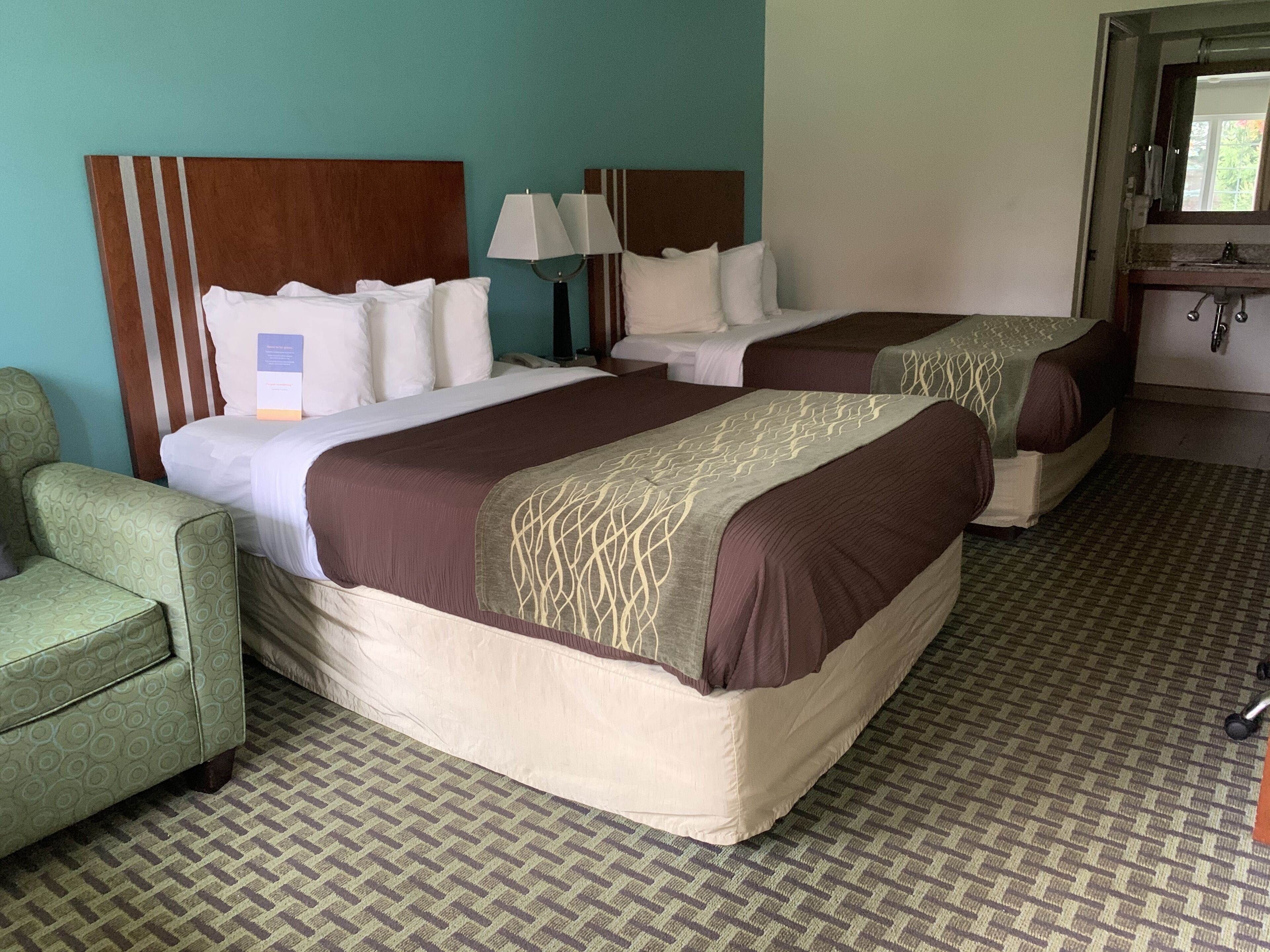 Habitación de hotel con dos camas, un sillón verde, un escritorio con lámpara y un baño visible a través de una puerta abierta.