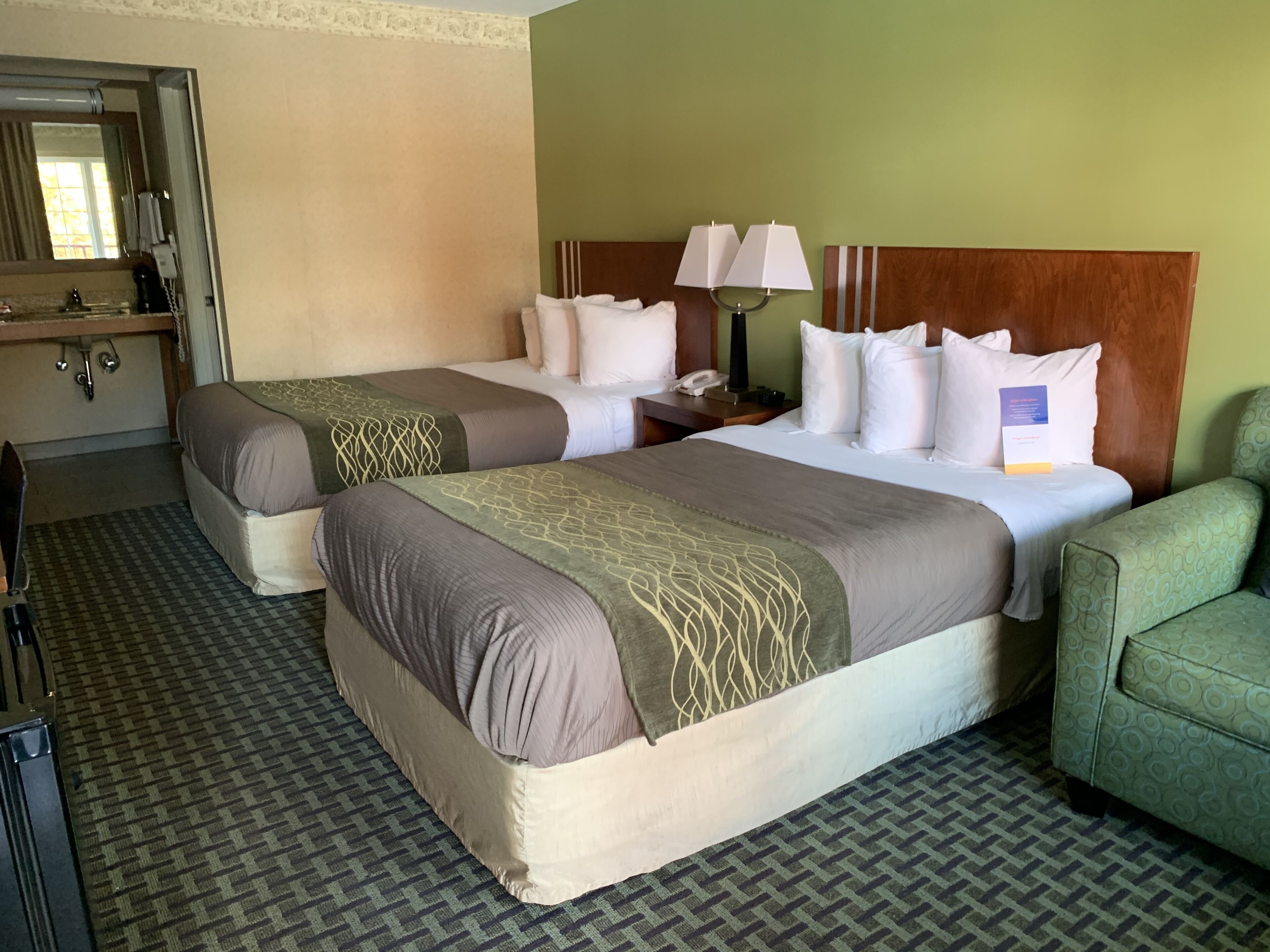 A hotel room with two beds, a green armchair, a desk with a lamp, and a mirror.