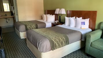 A hotel room with two beds, a green armchair, a desk with a lamp, and a mirror.