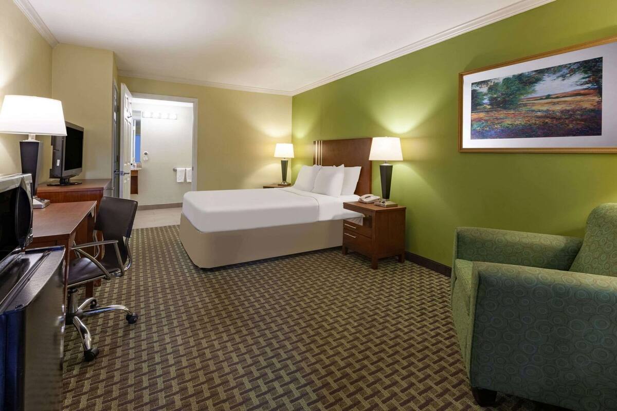 A hotel room with a bed, desk, chair, lamps, and a painting on the wall.