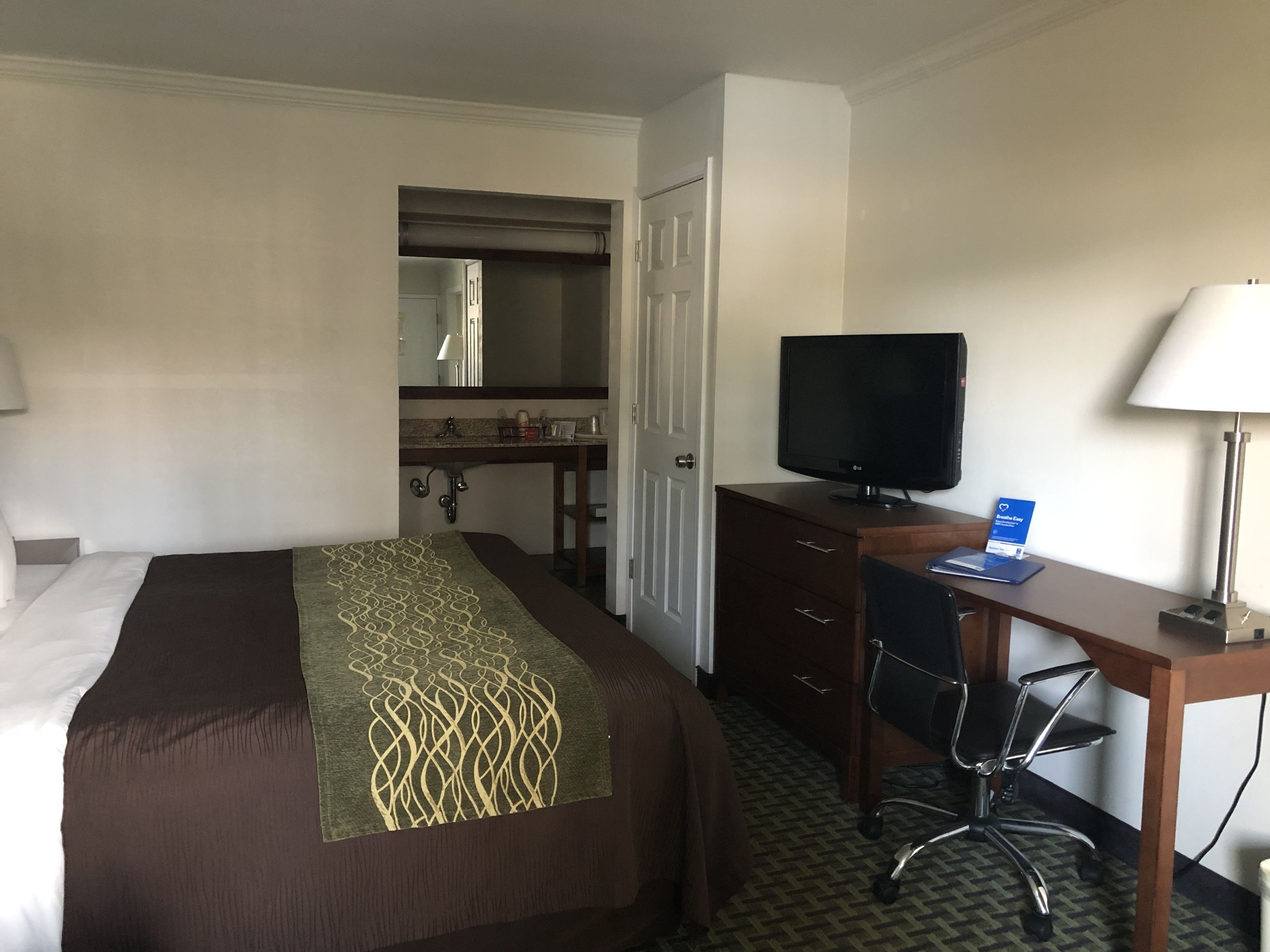 Une chambre d’hôtel avec un lit, un bureau avec un ordinateur, une télévision, une chaise, une lampe de bureau et une salle de bain visible à travers une porte ouverte.