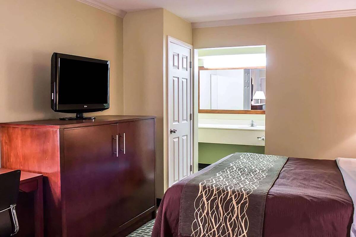 A hotel room with a bed, a TV on a wooden cabinet, a bathroom with a mirror, and a window.