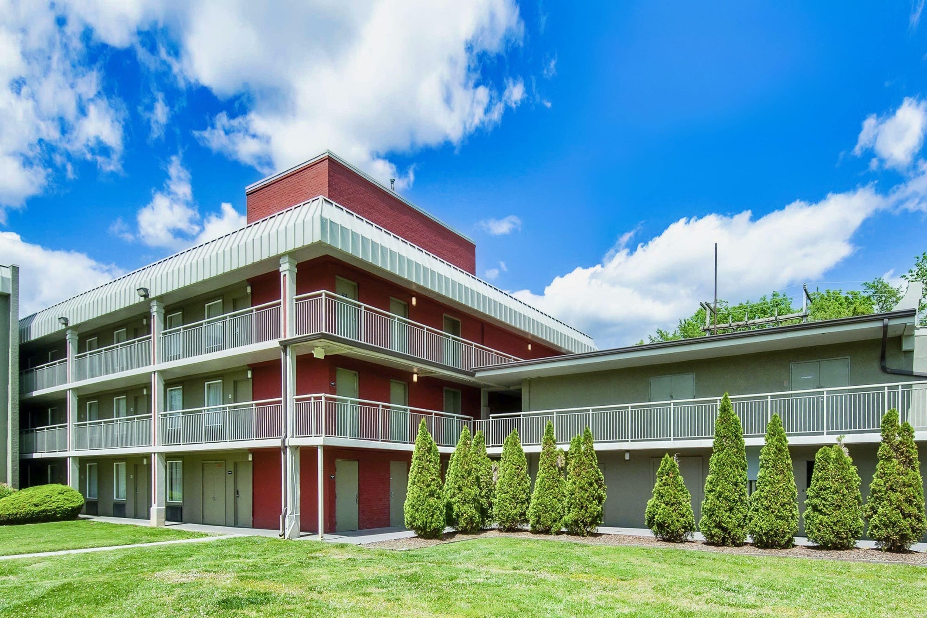 A multi-story building with balconies, a red and grey color scheme, and a well-maintained lawn with coniferous trees.