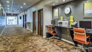 Un pasillo de hotel con alfombra estampada, un mostrador de recepción, dos sillas, un reloj y una impresora.