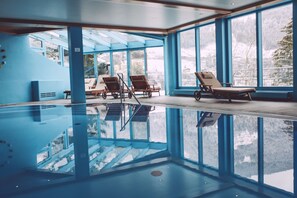Una gran piscina interior con paredes de vidrio transparente, sillones de madera y vistas a montañas nevadas.