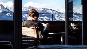 Una persona leyendo un libro junto a una ventana con vistas a montañas nevadas.