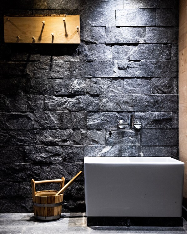 A bathroom with a stone wall, a wooden shelf with hooks, a white bathtub, and a wooden bucket with a brush. - Alpine Spa Hotel Haus Hirt (Bad Gastein)
