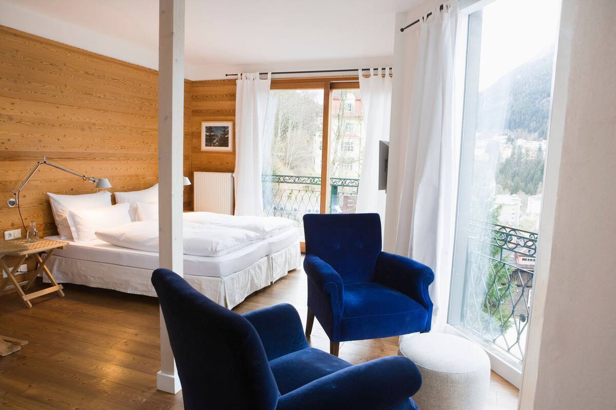 Un dormitorio con una cama, un sillón azul, una mesita de noche con una lámpara y una ventana que da a un balcón y edificios.