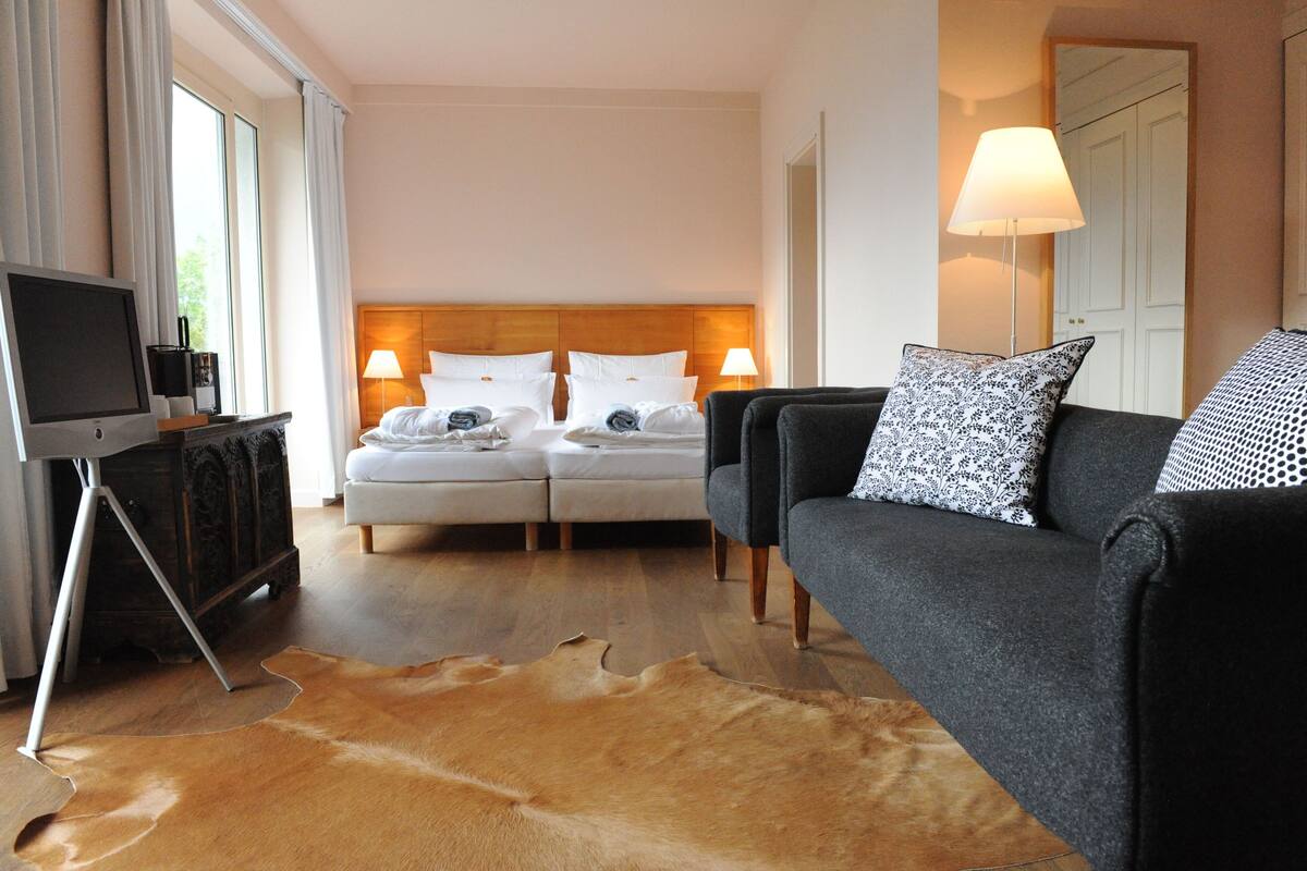 Habitación de hotel con cama, dos lámparas de noche, televisor sobre un mueble bajo, un sofá y un tapete grande de cuero.