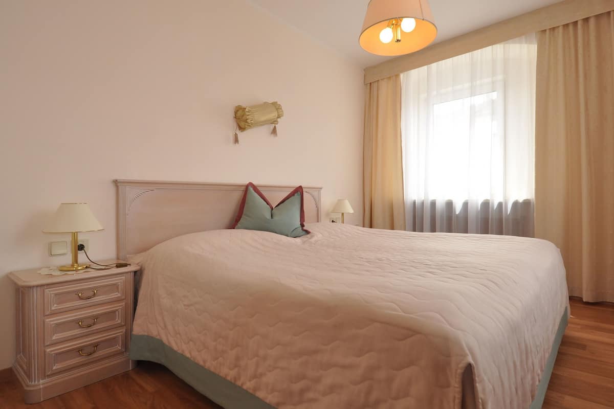 A neatly made bed with a quilted cover, a bedside table with a lamp, and a window with curtains.