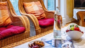 A hotel room with wicker chairs, a table with a bowl of fruit, a bottle of water, and a magazine.