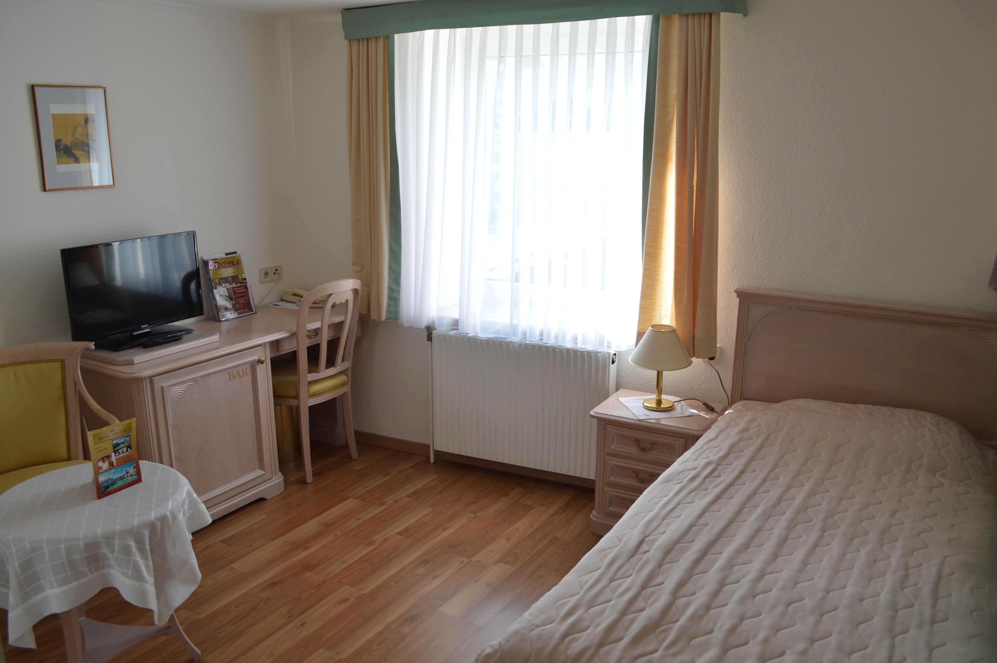 Quarto de hotel com cama, escrivaninha com televisão, cadeira, mesa com um livro e janela com cortinas.