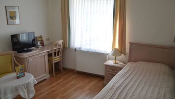 Quarto de hotel com cama, escrivaninha com televisão, cadeira, mesa com um livro e janela com cortinas.