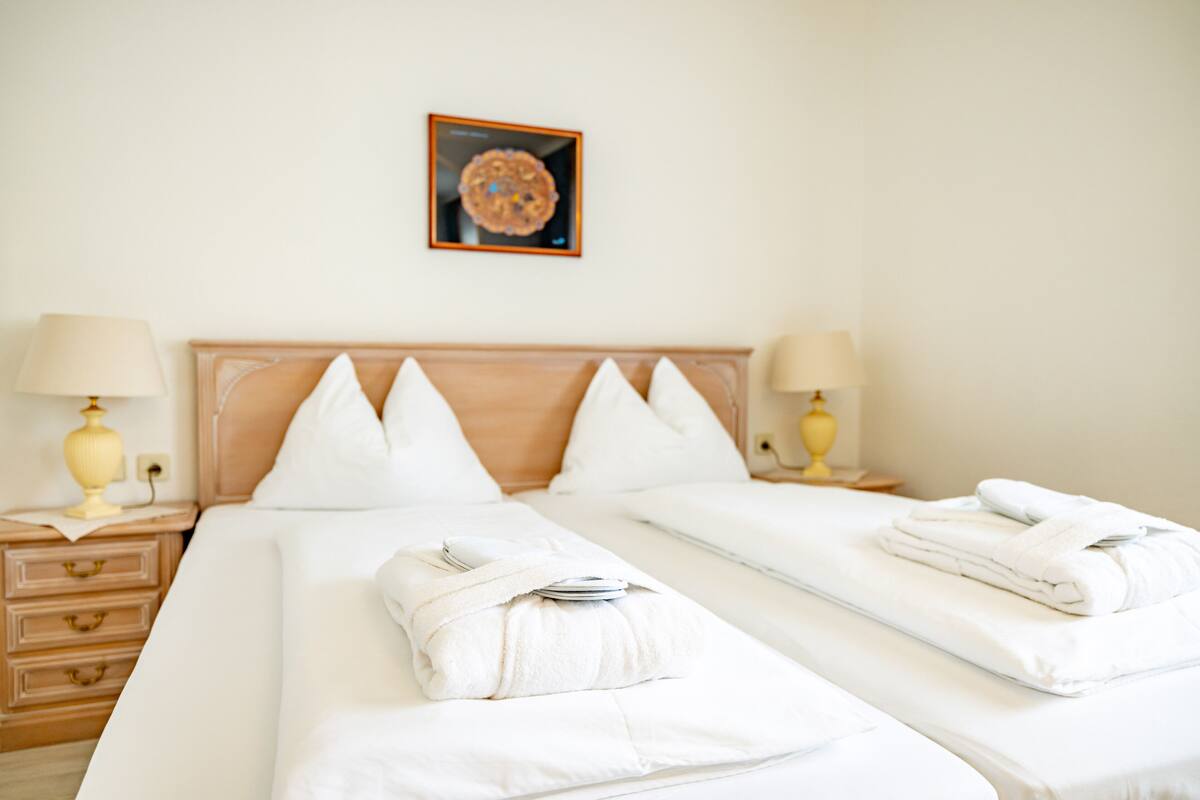 A hotel room with two beds, each with white linens and towels, a wooden headboard, bedside lamps, and a framed picture on the wall.