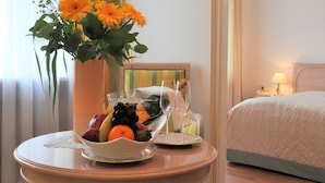 A hotel room with a bed, a bedside table with a vase of flowers, a fruit bowl, and a glass of water.