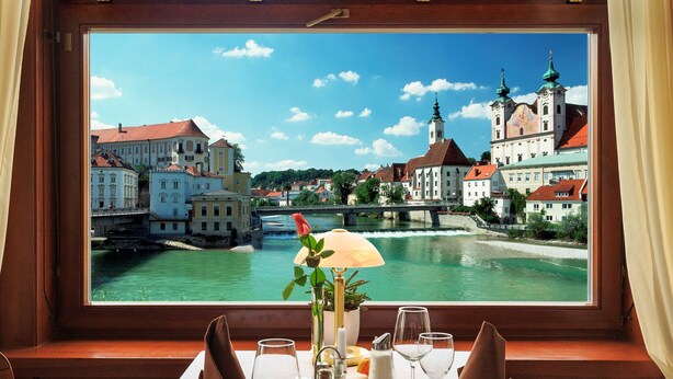 A dining table set with plates, glasses, and a flower vase, overlooking a river with buildings and a bridge.