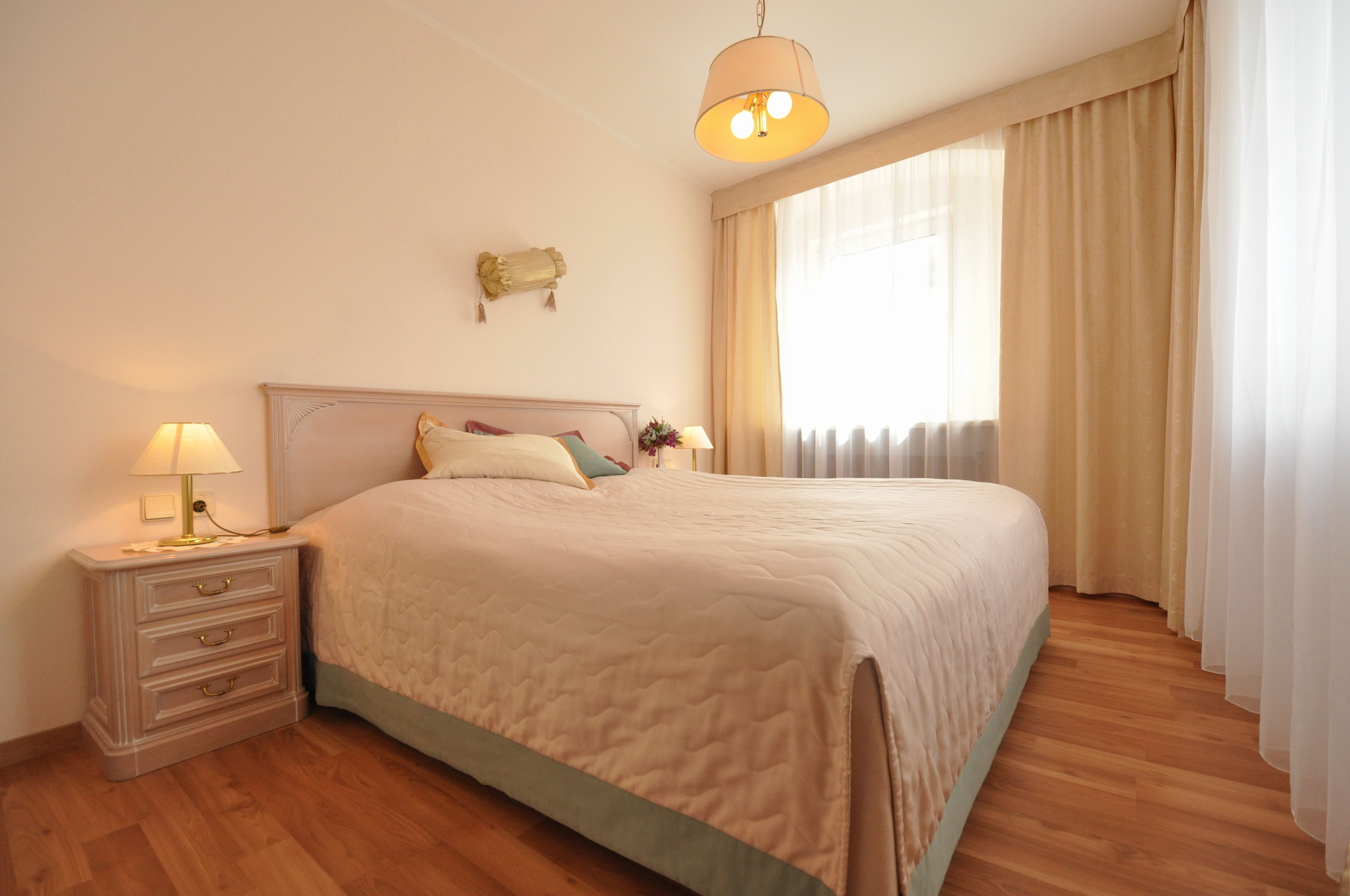 A neatly made bed with a headboard, a bedside table with a lamp, and a window with curtains.