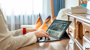 A person using a laptop on a wooden table in a room with a bed and a dresser.