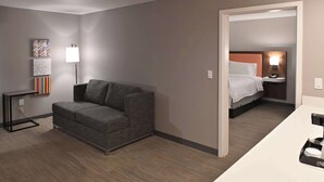 A hotel room with a gray sofa, a bedside table with a lamp, a bed with white linens, and a small nightstand with a phone. - Hampton Inn Fremont (Fremont)