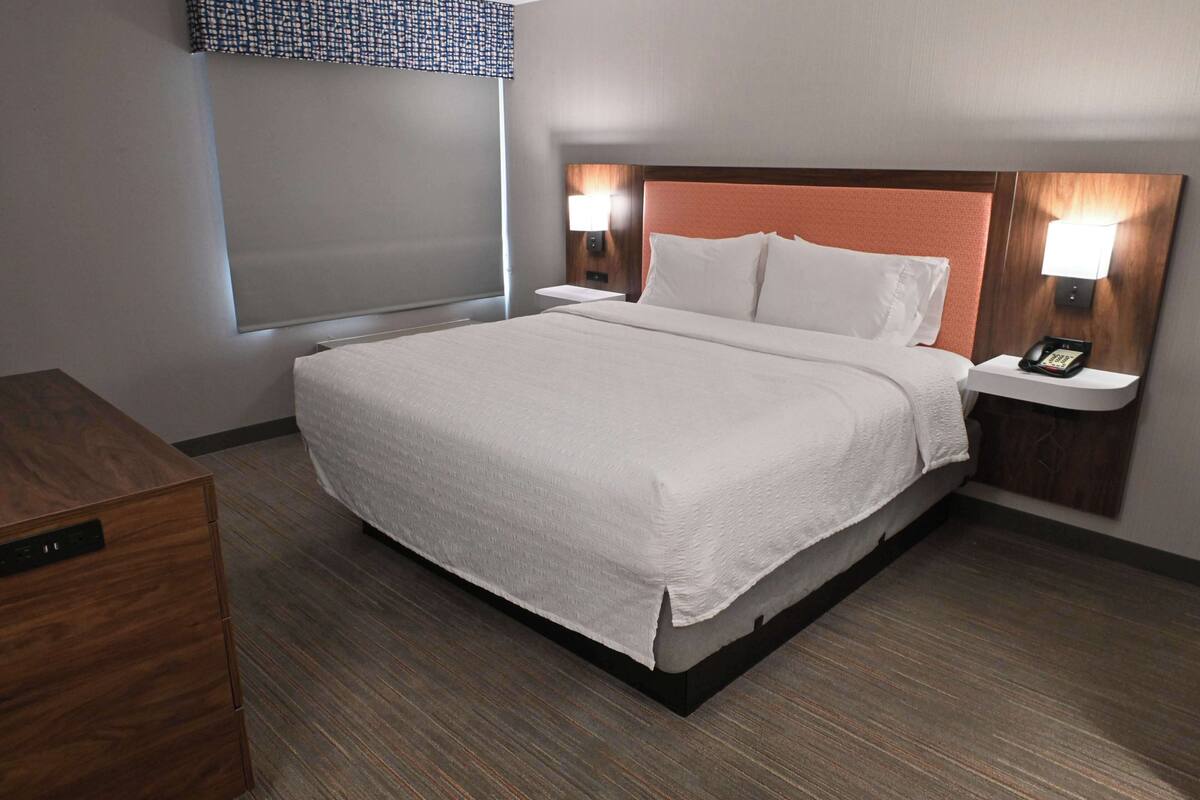 A hotel room with a large bed, two bedside tables with lamps, a wooden nightstand, and a window with blinds.