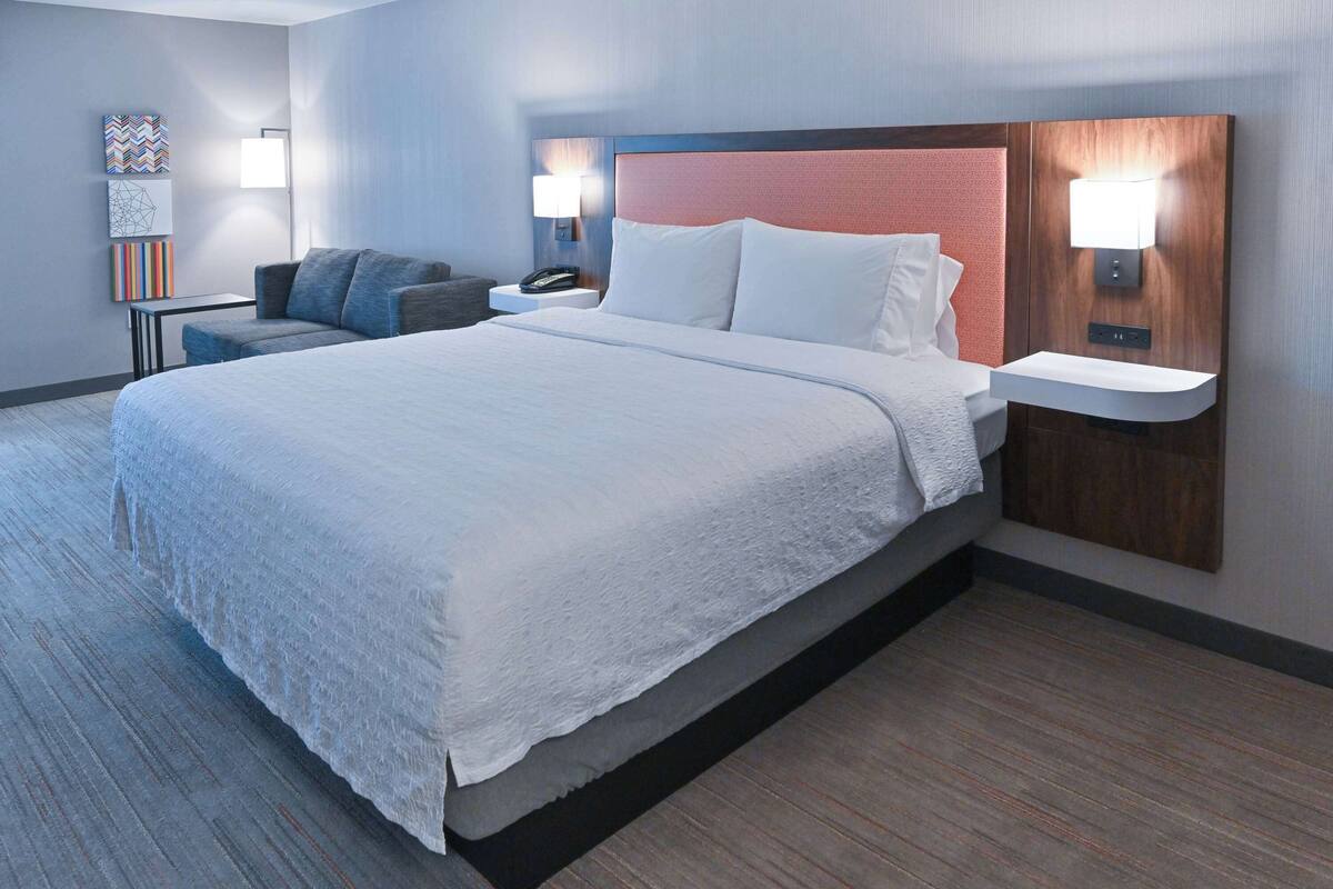 A modern hotel room with a large bed, a bedside table with a lamp, a small seating area with a chair, and artwork on the wall.