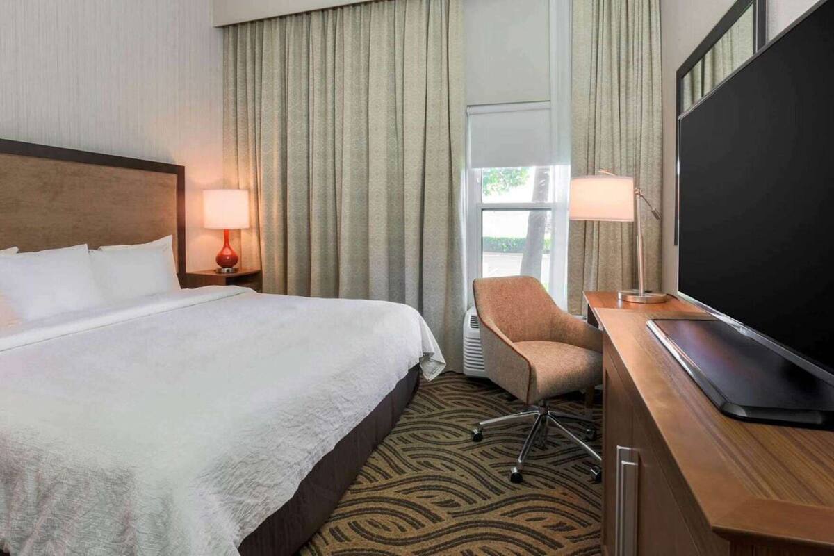 Ett hotellrum med en säng, ett skrivbord med en platt-TV, en stol och ett fönster med gardiner.