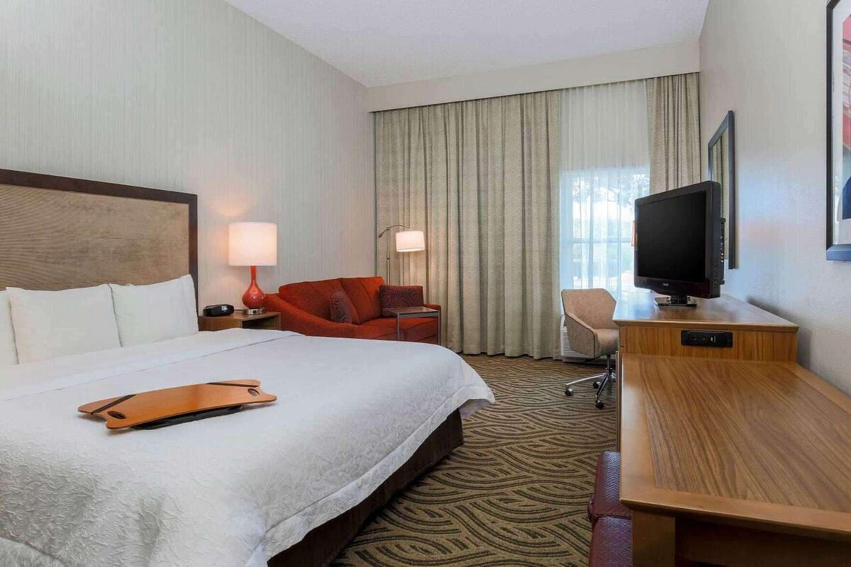 A hotel room with a bed, a desk, a television, a chair, and a large window with curtains.