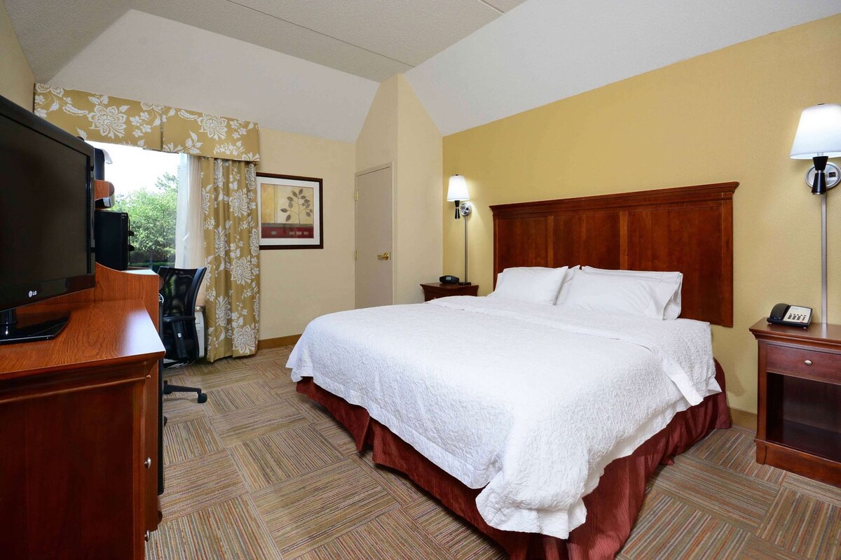 A hotel room with a bed, a desk with a TV, a chair, a lamp, and a telephone.