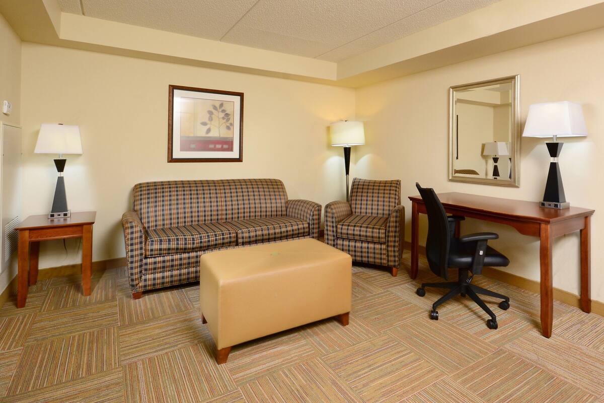 A hotel room with a plaid sofa, a matching armchair, a wooden desk, and a small ottoman. There are two lamps on side tables and a framed picture on the wall.