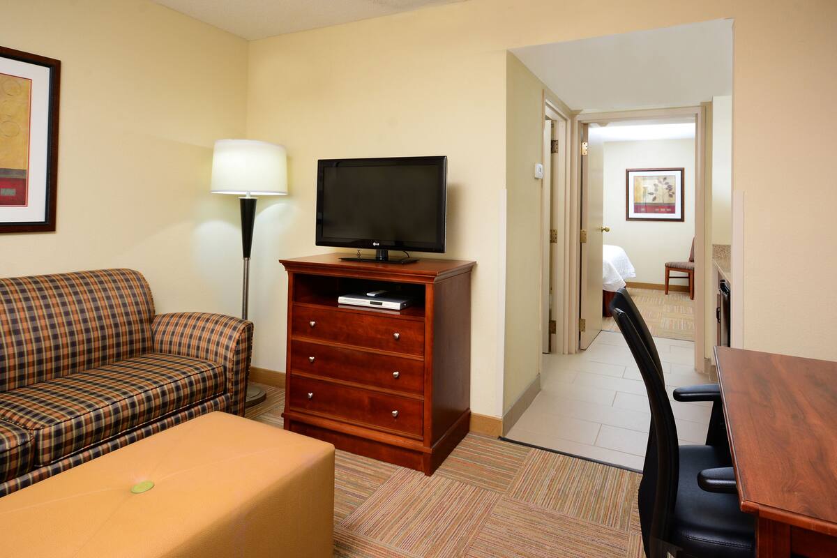 A hotel room with a plaid sofa, a wooden TV stand, a lamp, a small table, and a view of the hallway.