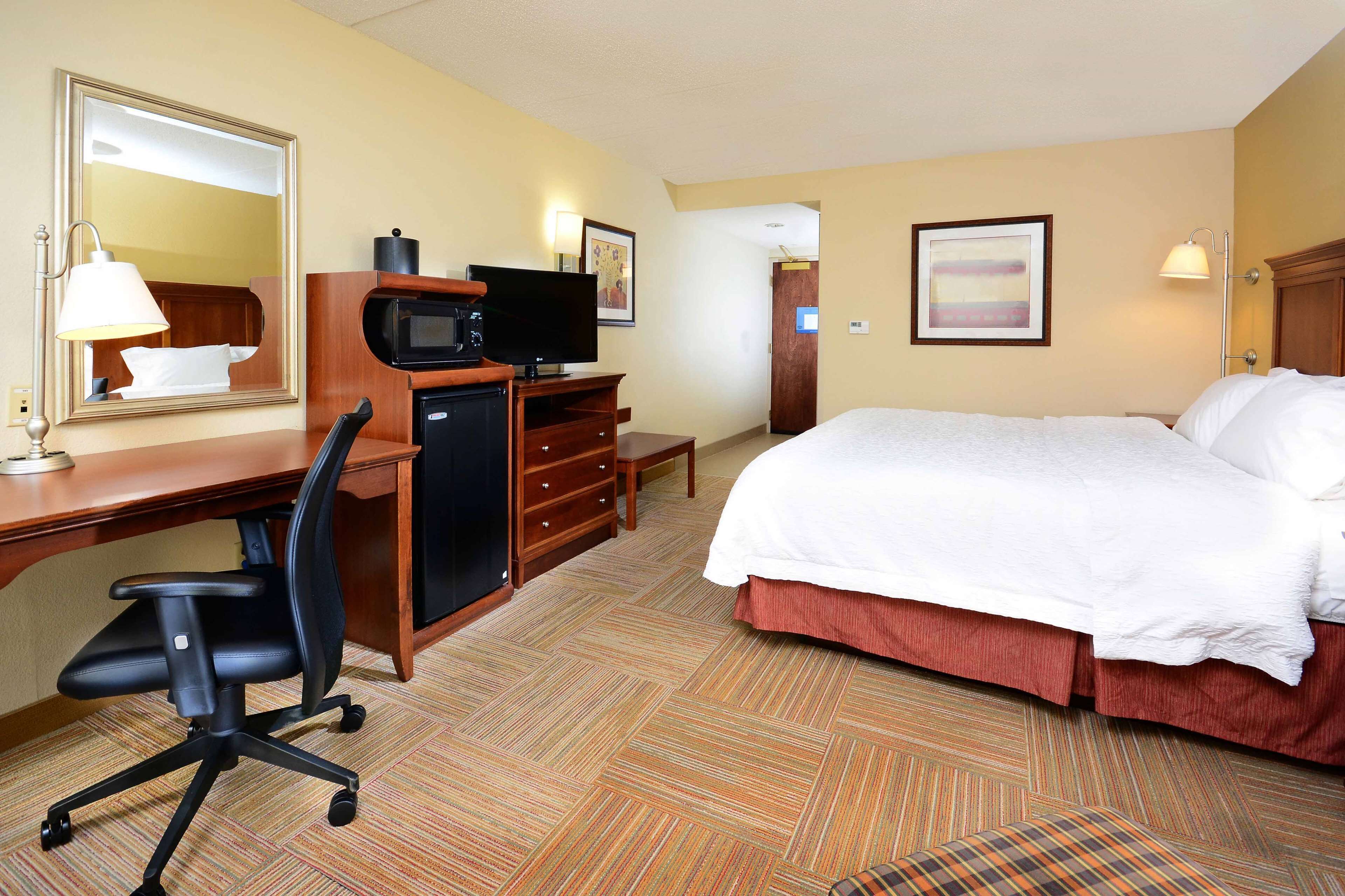 Quarto de hotel com cama, escrivaninha, cadeira, televisão e espelho.